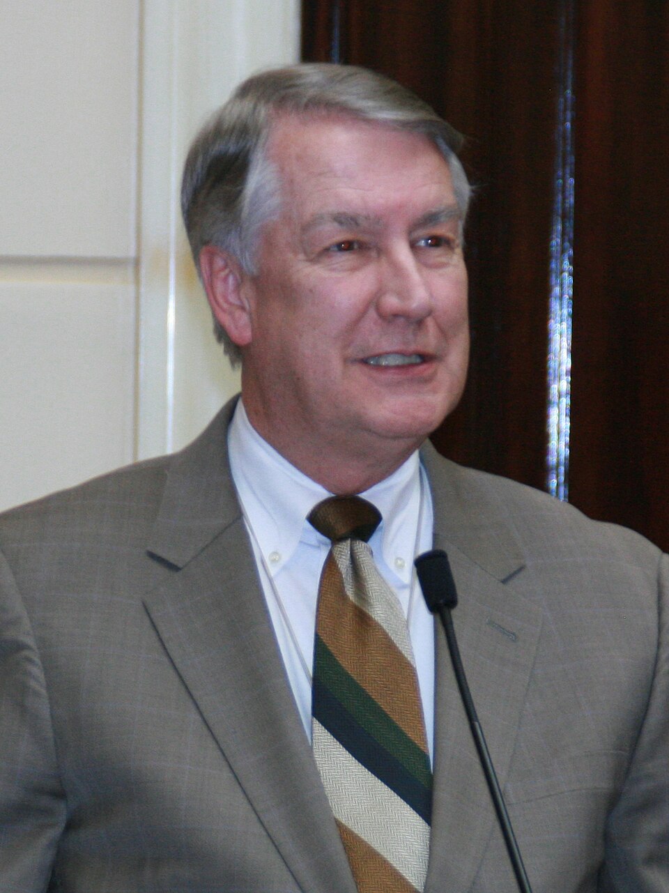 John Valentine, wearing a tan jacket and patterned tie, stands beside a microphone.